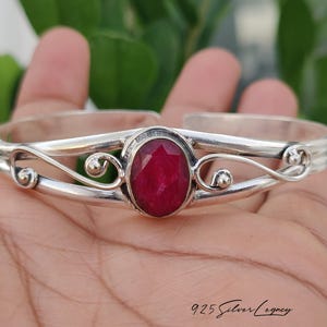May include: A silver cuff bracelet featuring a central oval-shaped faceted deep red gemstone. The metal band displays a polished finish with decorative swirling wire details on both sides of the stone setting. The text 925 Silver Legacy is visible.