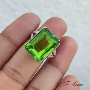 May include: A silver ring featuring a large, rectangular, bright green gemstone. The gemstone is set in a silver band. The ring is held in a hand, with the focus on the jewelry. The ring is a statement piece.