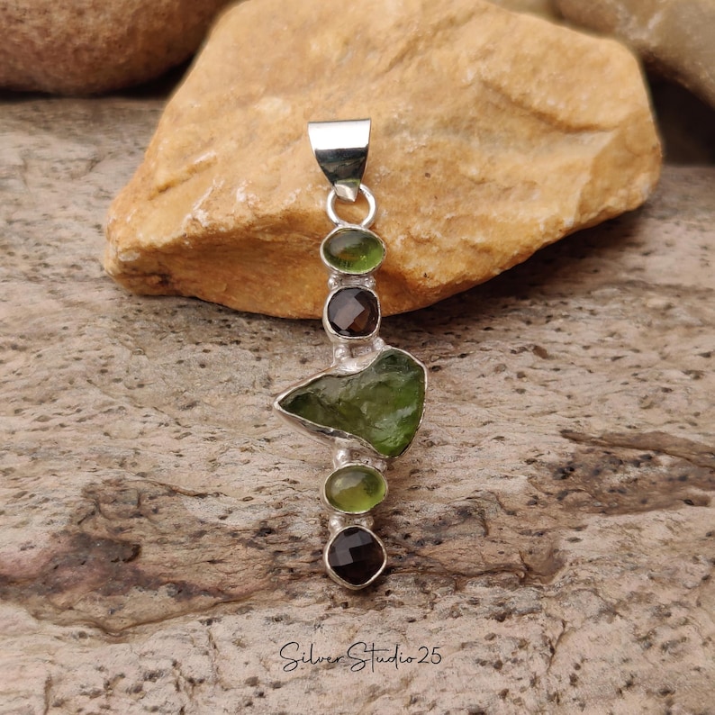 May include: A silver pendant featuring a series of green and brown gemstones. The pendant has a rectangular shape with a loop at the top for a chain. The gemstones are oval and irregular shapes. The text "Silver Studio 25" is at the bottom.
