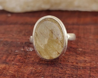 May include: A silver ring featuring an oval-shaped gemstone with golden inclusions. The ring's setting is a simple, polished silver band, and the gemstone has a translucent quality. The ring is set against a wooden surface.