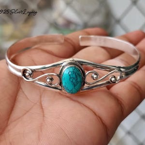May include: A silver cuff bracelet featuring a central oval turquoise stone. The bracelet is adorned with decorative swirls and small silver balls. The text "925 Silver Legacy" is visible in the upper left corner.