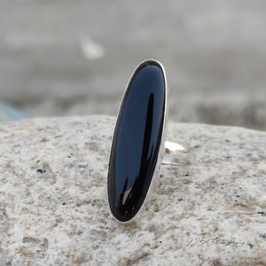 May include: A silver ring featuring a large, elongated black onyx stone. The polished black stone is set in a sleek, minimalist silver band. The ring is displayed on a textured, light-colored stone surface.