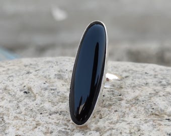 May include: A silver ring featuring a large, elongated black onyx stone. The polished black stone is set in a sleek, minimalist silver band. The ring is displayed on a textured, light-colored stone surface.