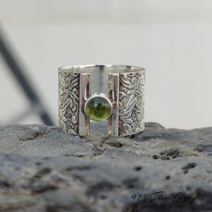 May include: A wide, sterling silver ring with an engraved floral pattern. The ring features a central, round, olive-green gemstone. The ring's design includes two vertical silver bars framing the gemstone. The ring is set on a dark grey stone.