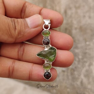 May include: A silver pendant featuring a raw, olive-green gemstone and four smaller, round gemstones in shades of green and brown. The pendant is held in a hand and has the text "Silver Studio 25" at the bottom.
