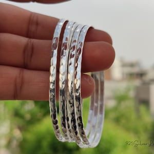 May include: A set of five hammered silver bangles. The bangles have a textured surface and are stacked together. The background is blurred, with green foliage and a building visible. The text "925 Silver Legacy" is in the bottom right corner.