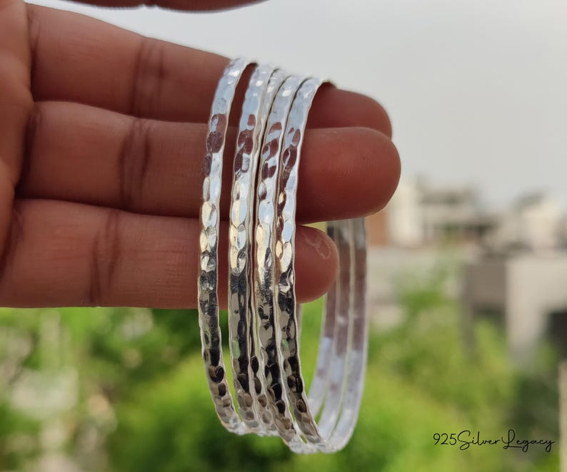 May include: A set of five hammered silver bangles. The bangles have a textured surface and are stacked together. The background is blurred, with green foliage and a building visible. The text "925 Silver Legacy" is in the bottom right corner.