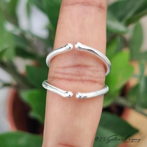 May include: Two polished silver open-ended rings with rounded, smooth terminals. The bands feature a simple, minimalist design with a reflective metallic finish. These adjustable rings are crafted from sterling silver, showcasing a clean, sleek aesthetic.