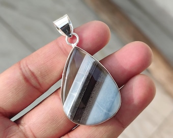 May include: A teardrop-shaped pendant with a striped pattern in shades of brown, white, and blue. The gemstone is set in a silver bezel with a silver bail. The pendant is held in a hand. The text "925 Silver Legacy" is at the bottom.