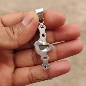 May include: A silver pendant with a unique, elongated design. The pendant features a central, triangular-shaped stone and several smaller, circular accents. The text "Silver Studio 25" is visible at the bottom. The pendant is held in a hand.