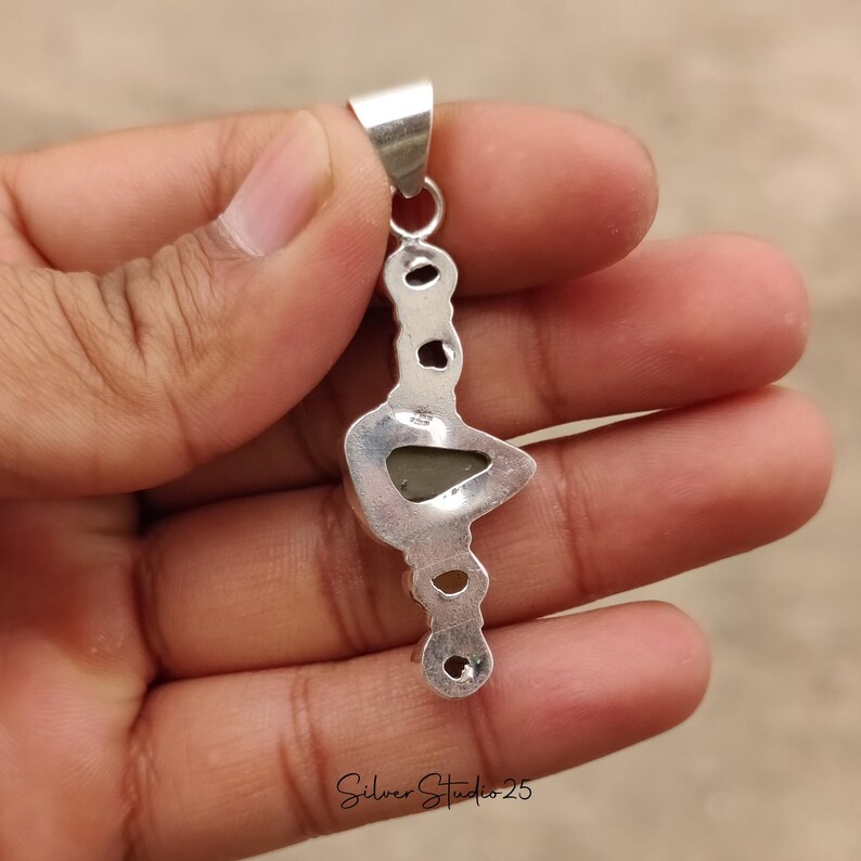 May include: A silver pendant with a unique, elongated design. The pendant features a central, triangular-shaped stone and several smaller, circular accents. The text "Silver Studio 25" is visible at the bottom. The pendant is held in a hand.