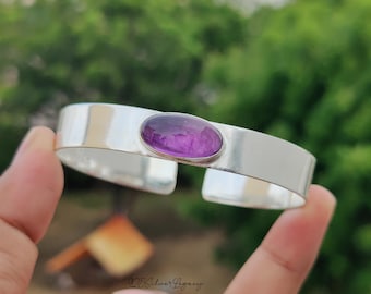 May include: A silver cuff bracelet featuring a large, oval, purple amethyst gemstone. The bracelet has a wide, polished band and a minimalist design. The gemstone is set in a silver bezel, adding a touch of elegance to the jewelry.