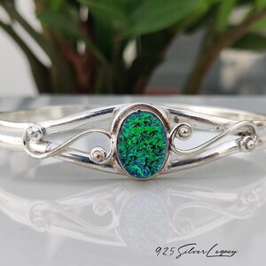 May include: A sterling silver cuff bracelet featuring a central oval-shaped iridescent green and blue stone. The metal band has a polished finish with decorative swirling scrollwork on both sides of the stone. The text 925 Silver Legacy is visible.