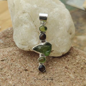 May include: A silver pendant featuring a large, irregular green gemstone, flanked by smaller green and brown stones. The pendant has a polished silver finish and is displayed on a light-colored rock. The text "Silver Studio 25" is visible.