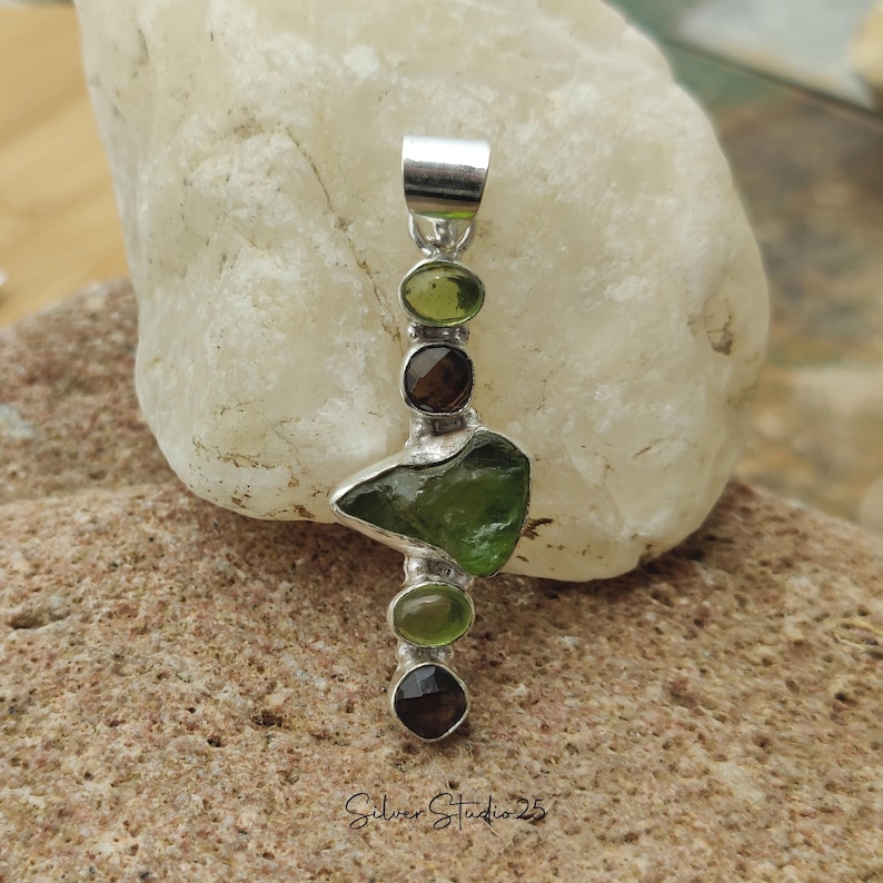 May include: A silver pendant featuring a large, irregular green gemstone, flanked by smaller green and brown stones. The pendant has a polished silver finish and is displayed on a light-colored rock. The text "Silver Studio 25" is visible.