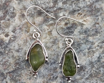 May include: A pair of silver earrings with a teardrop shape, each featuring a rough-cut, olive-green gemstone. The earrings have a simple hook design and are set against a textured gray background. The text "925 Silver Legacy" is visible at the bottom.