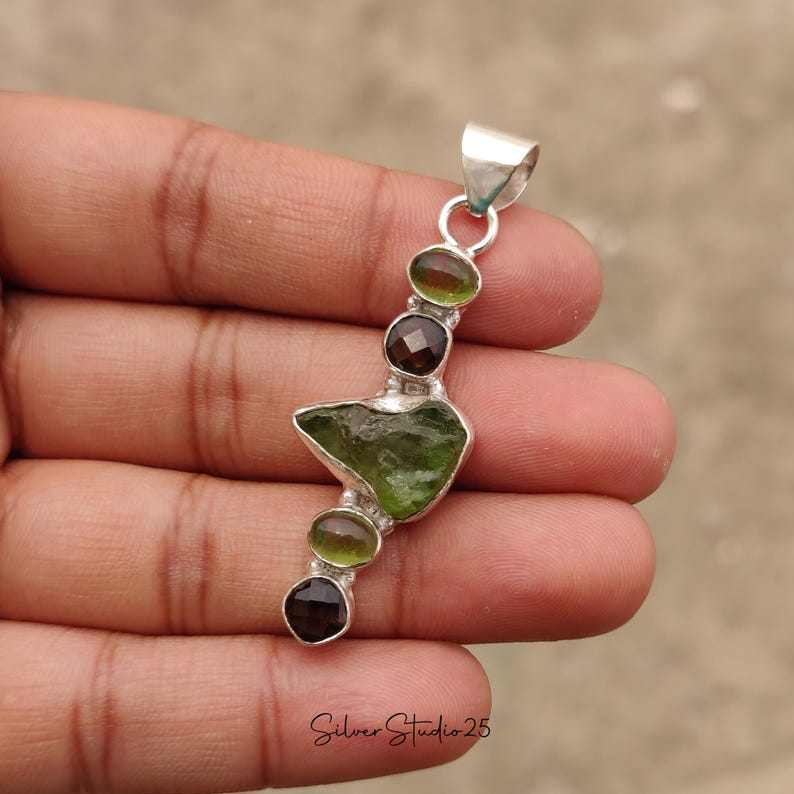 May include: A silver pendant featuring a series of gemstones. The pendant has a large, irregular green stone in the center, flanked by smaller, round green and brown stones. The text "Silver Studio 25" is at the bottom.