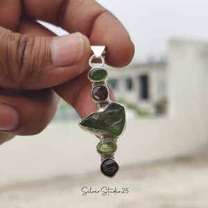 May include: A silver pendant featuring a series of green and brown gemstones. The pendant has a unique, organic shape with a large, irregularly shaped green stone at its center. The text "Silver Studio 25" is at the bottom.