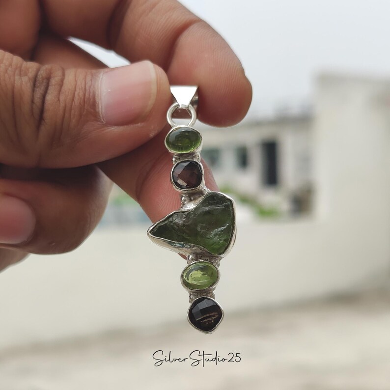 May include: A silver pendant featuring a series of green and brown gemstones. The pendant has a unique, organic shape with a large, irregularly shaped green stone at its center. The text "Silver Studio 25" is at the bottom.