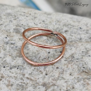 Puede incluir: Dos anillos de color cobre, uno ligeramente por encima del otro, creando un efecto en capas. Los anillos están colocados sobre una superficie de piedra gris claro moteada. Los anillos son simples y elegantes, con un diseño minimalista. El texto "925 Silver Legacy" está en la esquina superior derecha.
