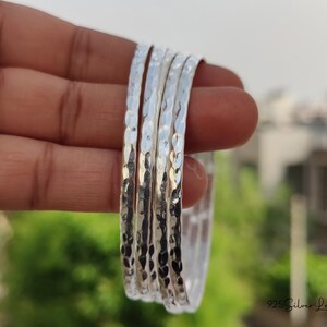 May include: A set of five silver bangles with a hammered texture. The bangles are held in a hand, with a blurred background of green foliage and a building. The bangles are made of a shiny, reflective metal.