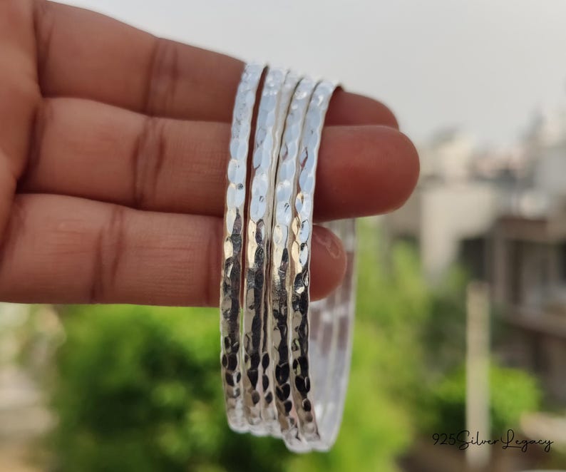 May include: A set of five silver bangles with a hammered texture. The bangles are held in a hand, with a blurred background of green foliage and a building. The bangles are made of a shiny, reflective metal.