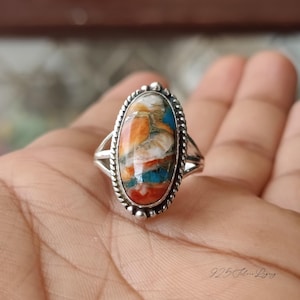 May include: A sterling silver ring with an oval-shaped, multicolored stone. The stone features a blend of orange, blue, white, and red hues. The ring has a detailed, beaded border and split shank design. The text "925 Silver Ligg" is visible.