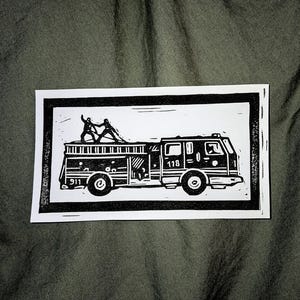 May include: Black and white linocut print of a fire engine with the numbers "118" and "911" visible. Two figures are on the roof of the engine. The print is on a white rectangular card with a black border.