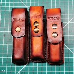 Peut inclure: Trois étuis en cuir marron avec le mot "KAOS" estampé sur le rabat. Chaque étui a une fermeture à bouton-pression avec du matériel de couleur laiton. Les étuis sont présentés sur un tapis de coupe vert.