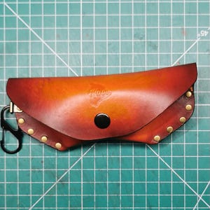 May include: A handcrafted leather glasses case in shades of brown and orange, secured with a black snap and brass rivets. The case features a black carabiner clip and the word "Hove" embossed on the front. The case is on a green cutting mat.