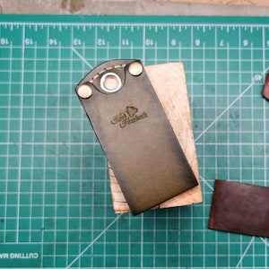 May include: A dark brown leather item with a metal grommet and rivets, resting on a wooden block. The item has the embossed text "King's Handcrafts". The background is a green cutting mat.
