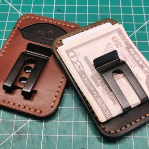 May include: Two leather wallets with metal money clips. One is brown, the other is black, and both have stitched edges. The black clip holds several banknotes. The wallets are on a green cutting mat.