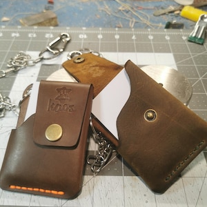 May include: Two brown leather cardholders with metal chain attachments. One is closed with a brass button, the other is open, revealing a white card. The closed cardholder has the word "kaos" printed on it.