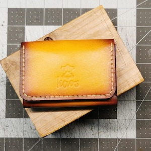 May include: A handcrafted leather wallet with a gradient yellow and brown finish. The wallet features stitched edges and a flap closure. The word "kaos" is embossed on the front. The wallet is resting on a wooden block.