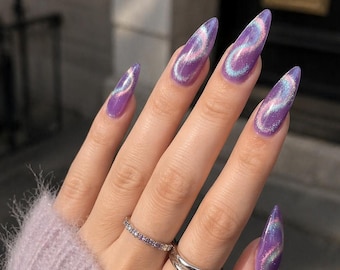 May include: Long almond-shaped nails with a purple cat-eye magnetic polish finish featuring a swirling iridescent shimmer. The fingers are adorned with two silver-toned rings, one thin band with small stones and one plain double-band ring.
