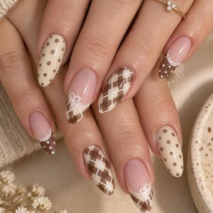 May include: Almond-shaped nails featuring a mix of designs including cream-colored nails with brown polka dots, brown and white plaid patterns, and French tips with white lace-like detailing. A thin gold ring with three small stones is worn.