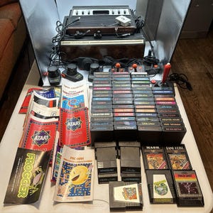 May include: A retro Atari video game console, controllers, and a selection of game cartridges and manuals. The console is brown and black. The game cartridges have colourful labels. The manuals are red and white. A nostalgic collection of gaming items.