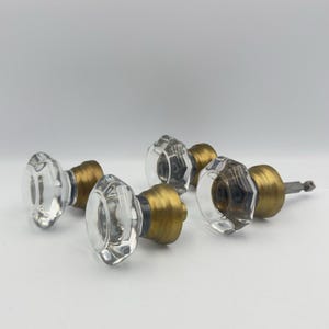 May include: Three door knobs featuring clear, faceted glass handles with an octagonal shape. Each knob is attached to a cylindrical, brushed brass-toned metal base. One knob shows a thin, square-shaped metal spindle extending from the back.