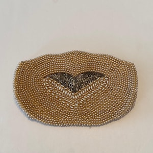 Puede incluir: Un bolso de mano vintage con cuentas de color oro claro, con un diseño en forma de corazón en el centro. El corazón está hecho con cuentas plateadas y oscuras, creando un contraste llamativo. El bolso tiene forma rectangular redondeada.