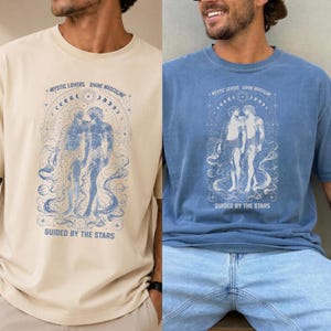 Könnte beinhalten: Zwei T-Shirts, eines in Beige und eines in Blau, jeweils mit einem himmlischen Design mit zwei Figuren und dem Text "MYSTIC LOVERS DIVINE MASCULINE" und "GUIDED BY THE STARS". Das Design ist hellblau.