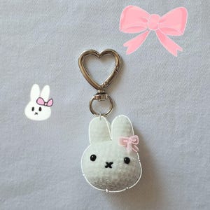 May include: A white bunny keychain with a pink bow, black eyes, and a stitched black mouth. The keychain has a silver heart-shaped clasp. A pink bow illustration is in the background.