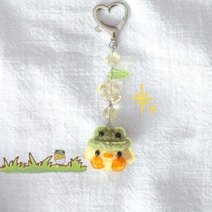May include: A keychain featuring a small, crocheted yellow chick wearing a green hat, suspended from a silver-toned heart-shaped clasp. The chain includes clear star-shaped, leaf-shaped, and round beads. A small graphic of a chick in a shell is visible.