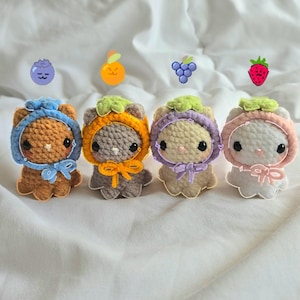May include: Four handmade crochet animal plushies, each with a different fruit-themed hood and bow. The plushies are brown, gray, beige, and white, with blue, orange, purple, and pink accents. Each plushie has a different fruit icon above it: blueberry, orange, grapes, and strawberry.