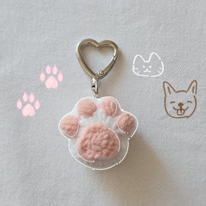 May include: A cat paw-shaped keychain with a heart-shaped clasp. The paw has a white base with pink pads and a clear outline. Pink paw prints and cat and dog sketches are in the background.