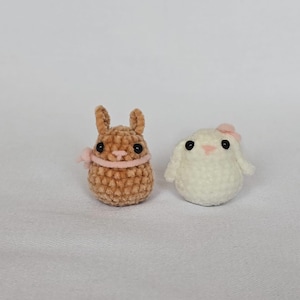 Op de afbeelding: Twee kleine, handgemaakte gehaakte konijntjes. E&eacute;n is bruin met een roze lint om de nek en de andere is wit met een roze strik. De konijntjes hebben zwarte ogen en zijn gemaakt van zacht, getextureerd garen. Ze staan op een witte ondergrond.