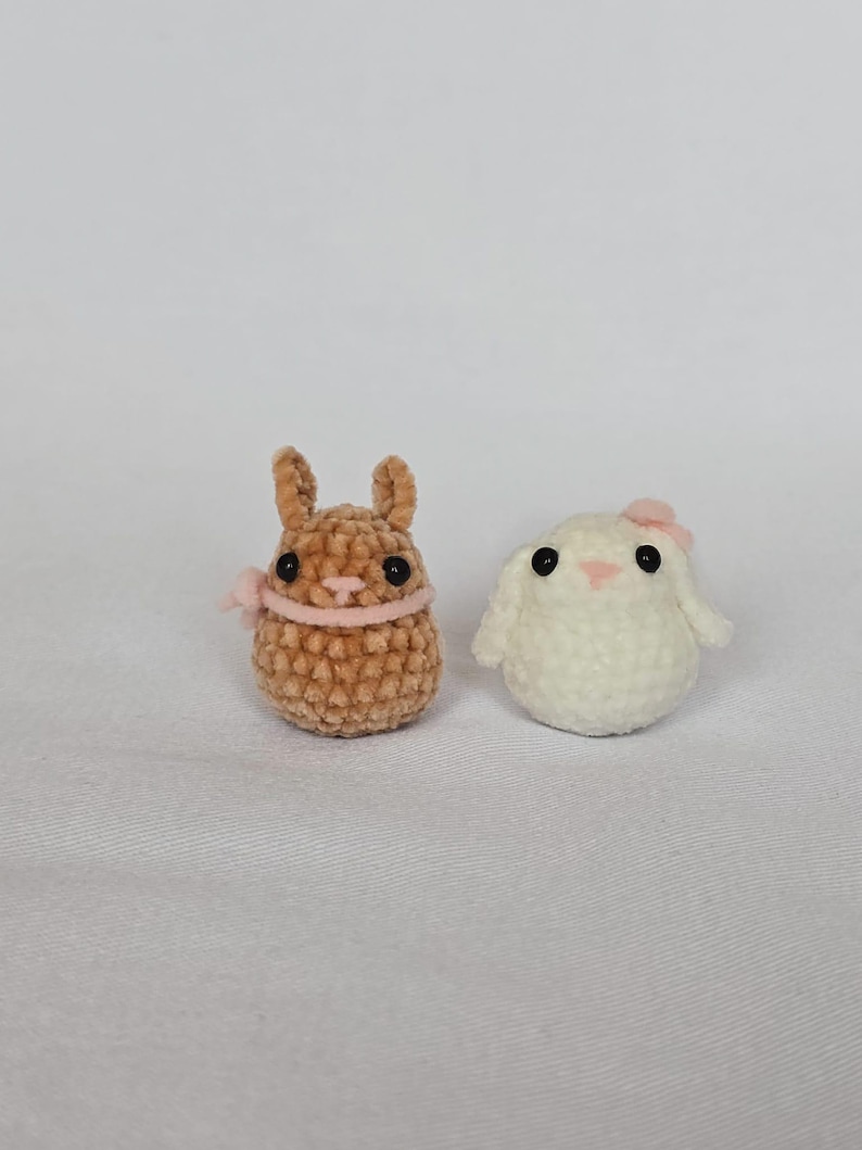 Op de afbeelding: Twee kleine, handgemaakte gehaakte konijntjes. E&eacute;n is bruin met een roze lint om de nek en de andere is wit met een roze strik. De konijntjes hebben zwarte ogen en zijn gemaakt van zacht, getextureerd garen. Ze staan op een witte ondergrond.