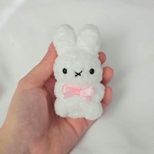 Puede incluir: Un pequeño conejito de peluche blanco con ojos negros y una 'X' cosida como boca. El conejito tiene un lazo rosa atado alrededor del cuello. El juguete está hecho de un material suave y difuso.