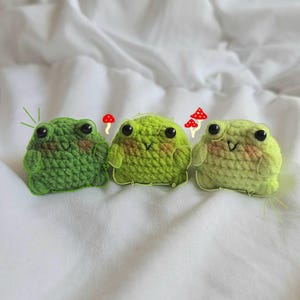 Può includere: Tre peluche a forma di rana fatti a mano all'uncinetto in varie tonalità di verde. Ogni rana ha occhi neri e una bocca sorridente. Piccoli accenti di funghi rossi e bianchi sono posti vicino alle rane.