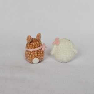 Op de afbeelding: Twee kleine, gehaakte dierenfiguren. E&eacute;n is bruin met een roze strik en een witte staart. De andere is wit met een roze strik. De figuren zijn waarschijnlijk handgemaakt en staan op een witte achtergrond.