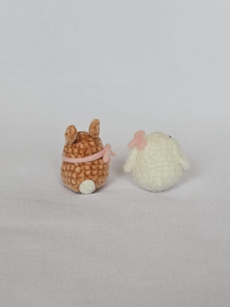 Op de afbeelding: Twee kleine, gehaakte dierenfiguren. E&eacute;n is bruin met een roze strik en een witte staart. De andere is wit met een roze strik. De figuren zijn waarschijnlijk handgemaakt en staan op een witte achtergrond.